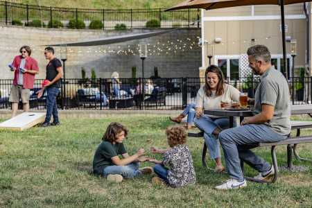 A young family enjoying food and drinks at a picnic table in Azalea Bar & Kitchen's lawn on a sunny summer afternoon. In the background, two men are playing cornhole.