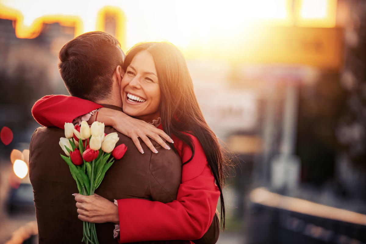 A dark-haired male-and-female couple hugs as the sun sets. The woman holds a bouquet of red and white flowers behind her male partner's back. 
