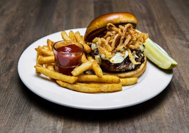 An All-In burger from Traditions on the Lake topped with fried onion and cheese and plated alongside thick-cut home fries and a pickle spear.