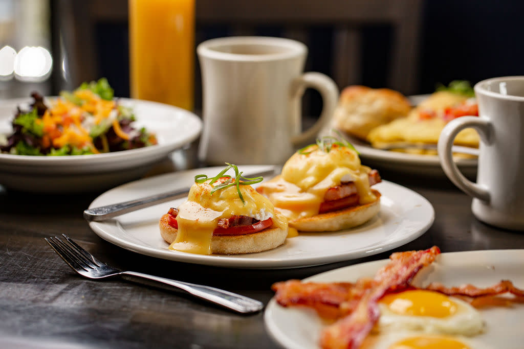 An order of eggs Benedict is accompanied by sunny side up eggs, crispy slices of bacon, and two mugs of freshly brewed coffee.