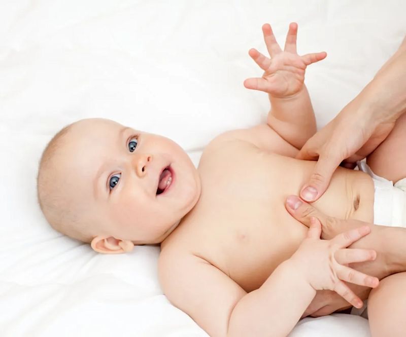 Smiling baby lying on a white bed while an parent's hands gently touch its belly.
