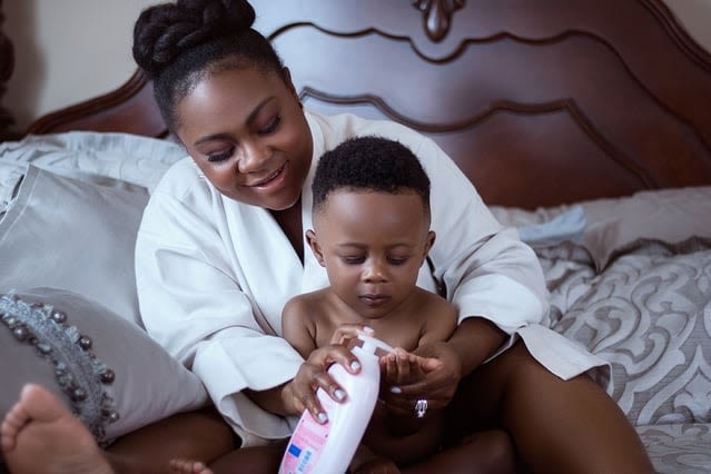 A mother helps her baby apply lotion while sitting on a bed with patterned pillows and a wooden headboard.