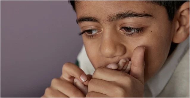 A child is thoughtfully biting their fingernail in a neutral-colored room, their hands close to their face, staring intently at something out of the frame.
