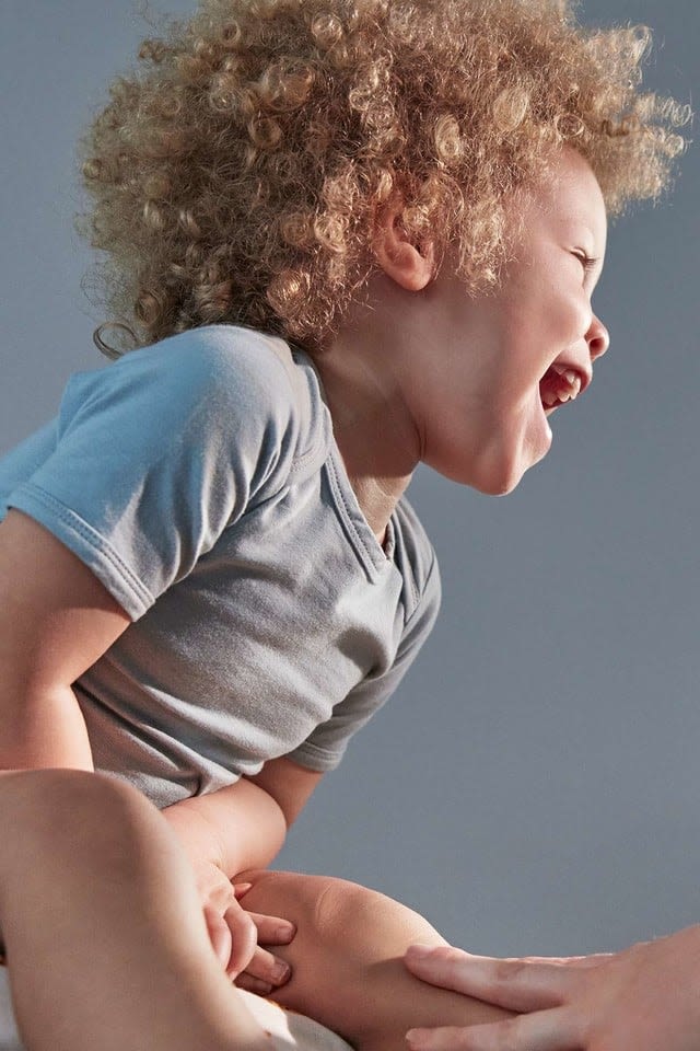 A toddler with curly hair, in a gray shirt, is laughing and looking to the right, while holding an adult's hand, in a well-lit room.