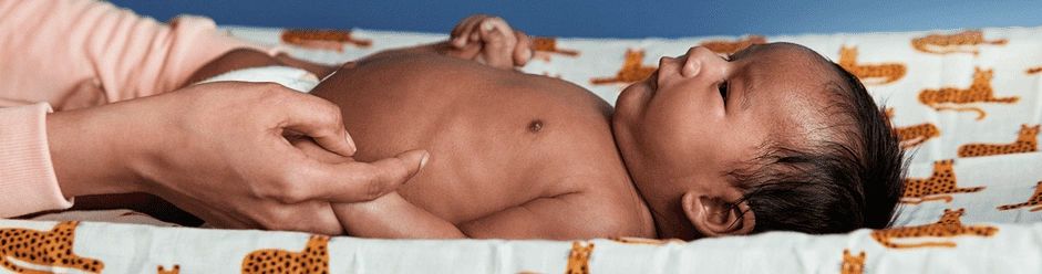 A parent holds a baby's hand while the baby lies on a cheetah-print changing pad, wearing a diaper and looking up.