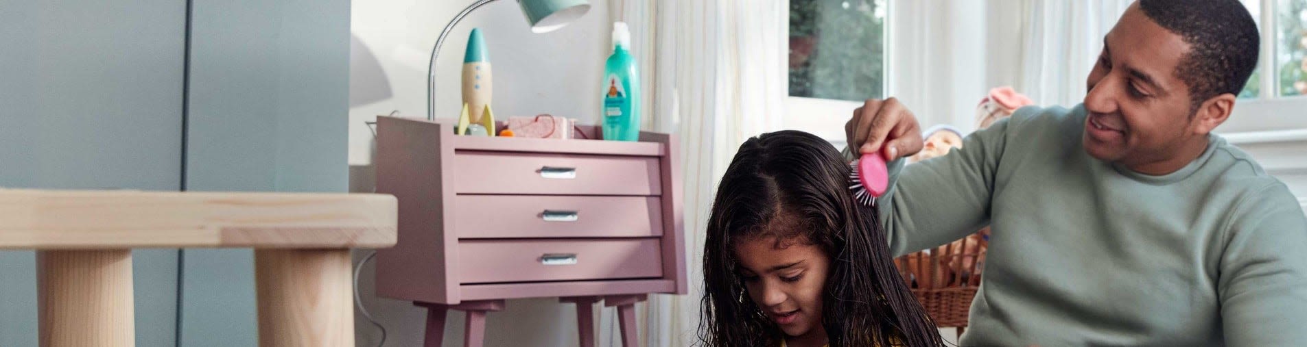 A smiling man brushes the wet hair of a seated girl in a bright room with a pink dresser and window in the background.