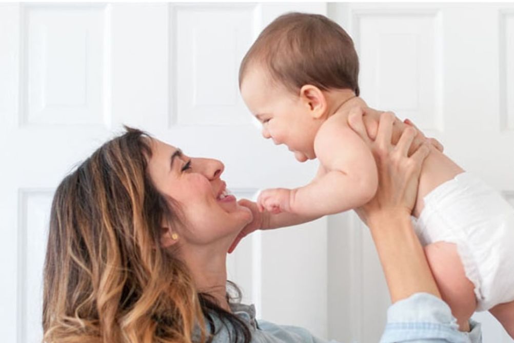 A mother is lifting a smiling baby in the air, both looking at each other and smiling joyfully.