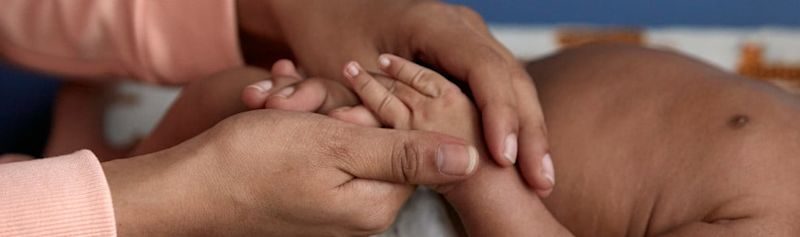Adult hands gently holding a baby's hands; context includes a close-up of the baby's torso laying on a blanket.