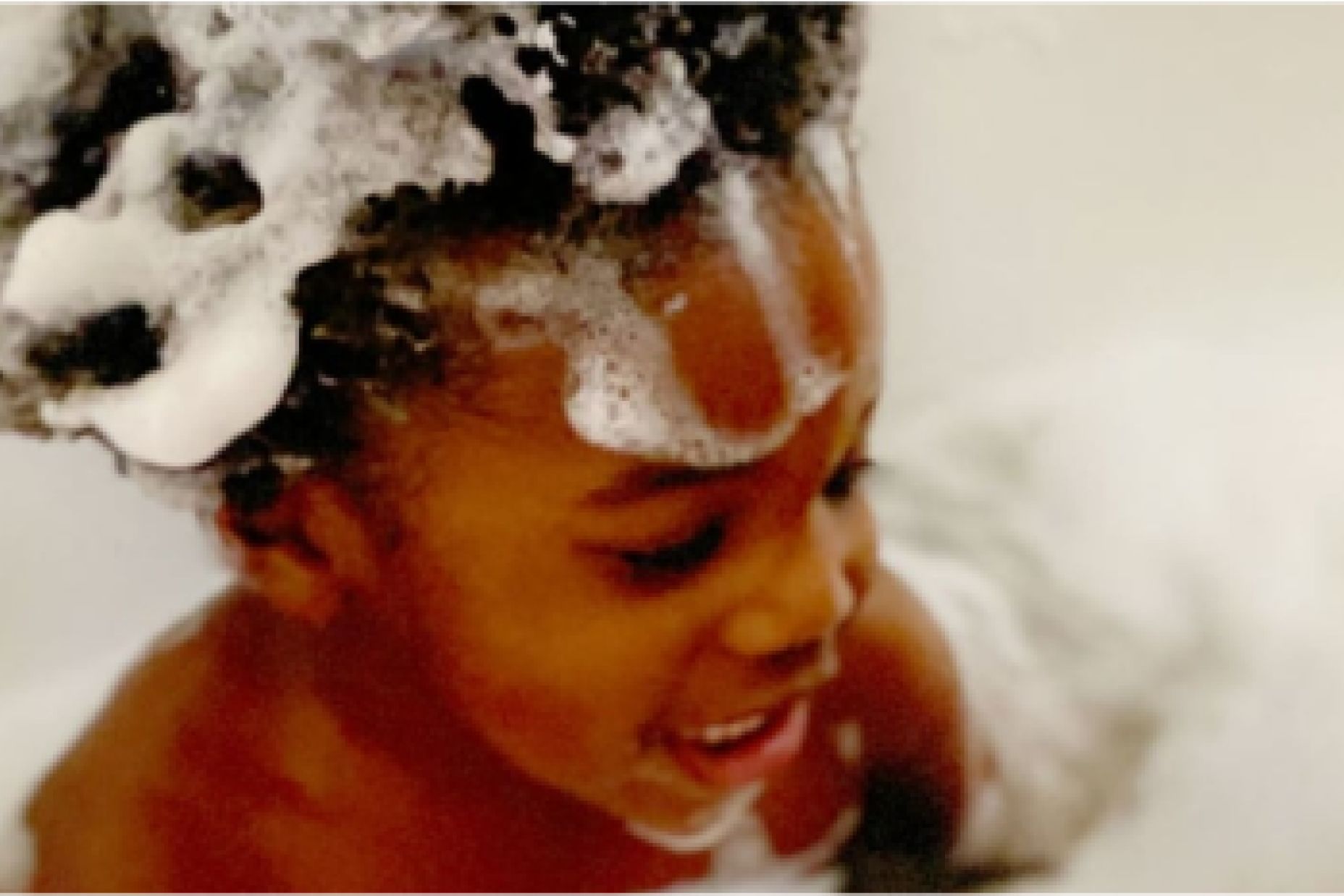 Child with curly hair smiles while enjoying a bath, covered in soapy bubbles.