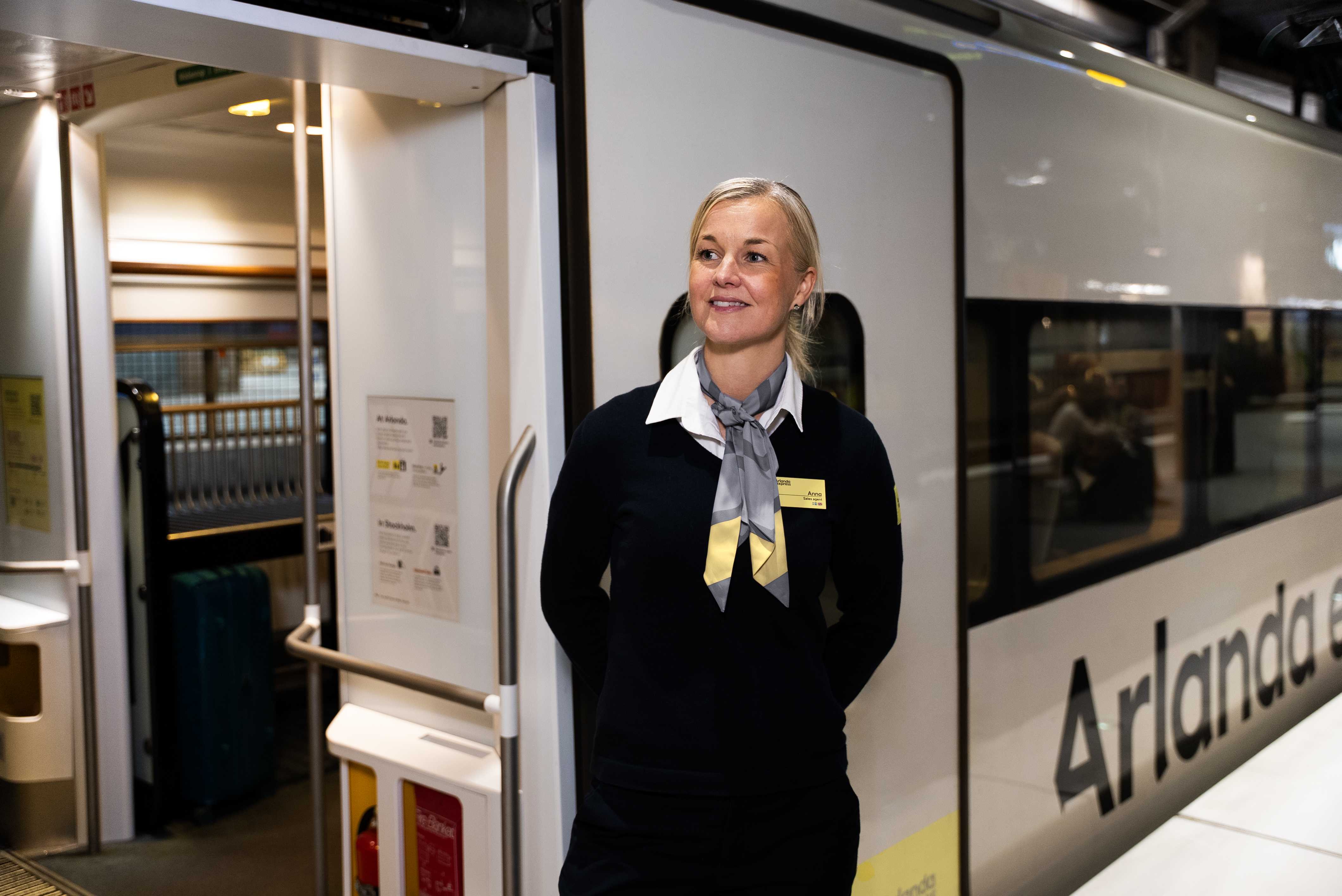 A friendly Arlanda express train attendant standing confidently on the platform at Arlanda Airport next to the sleek yellow and white train, ready to assist departing passengers.