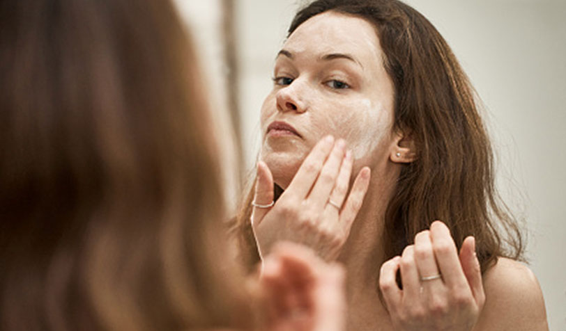 Image shows a person applying a white facial cleanser or cream to their face in front of a mirror