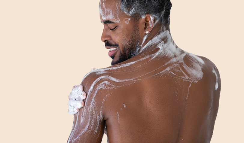 A smiling man spreading foaming body wash over his shoulder on a beige background