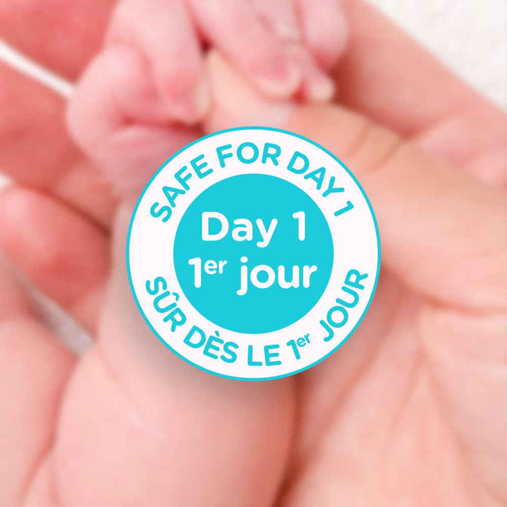 Aveeno® Baby product claim indicating 'Safe For Day One' and close-up of parent's hand holding their newborn baby's hand in the background