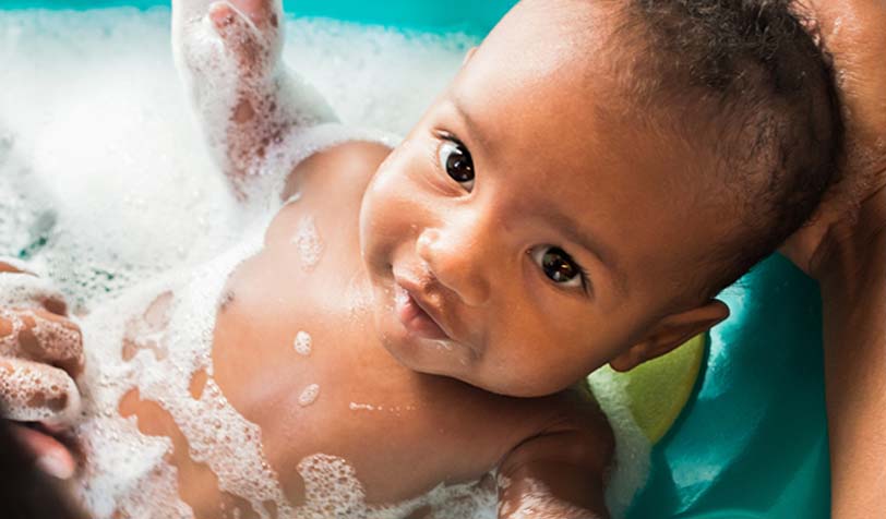 A smiling baby in a baby bath tub, surrounded by bubbles and water