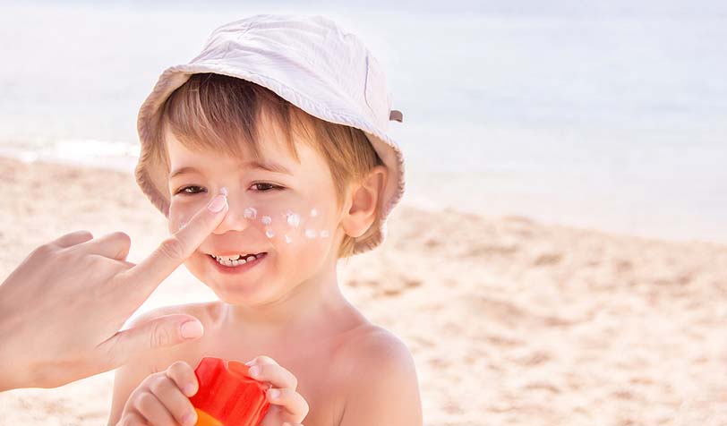 A smiling child wearing a white bucket hat getting sunscreen applied to his face at the beach on a sunny day