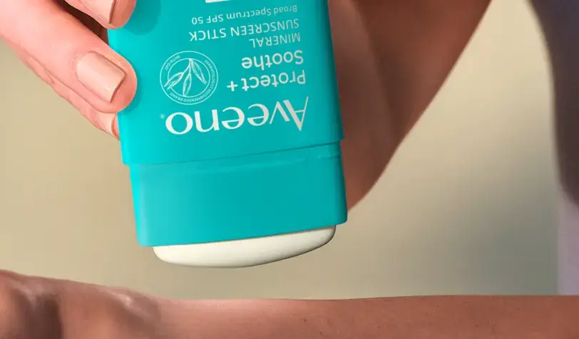 Hand applying a turquoise Aveeno Protect + Soothe Mineral Sunscreen Stick SPF 50 to a forearm