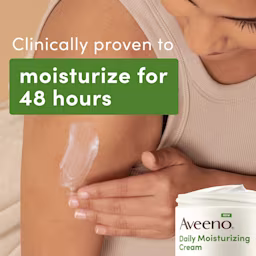 A woman wearing a beige tank top applies daily moisturizing cream to her arm. The cream is white and she is using her left hand to apply it. The background is a neutral beige color. The image says, "Clinically proven to moisturize for 48 hours."