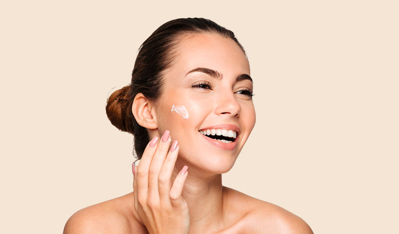 A woman smiles as she gently applies cream to her face, showcasing a moment of self-care