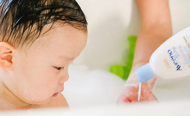 Person dispensing Aveeno® Baby Cleansing Therapy next to a baby sitting in a bathtub full of bubbles