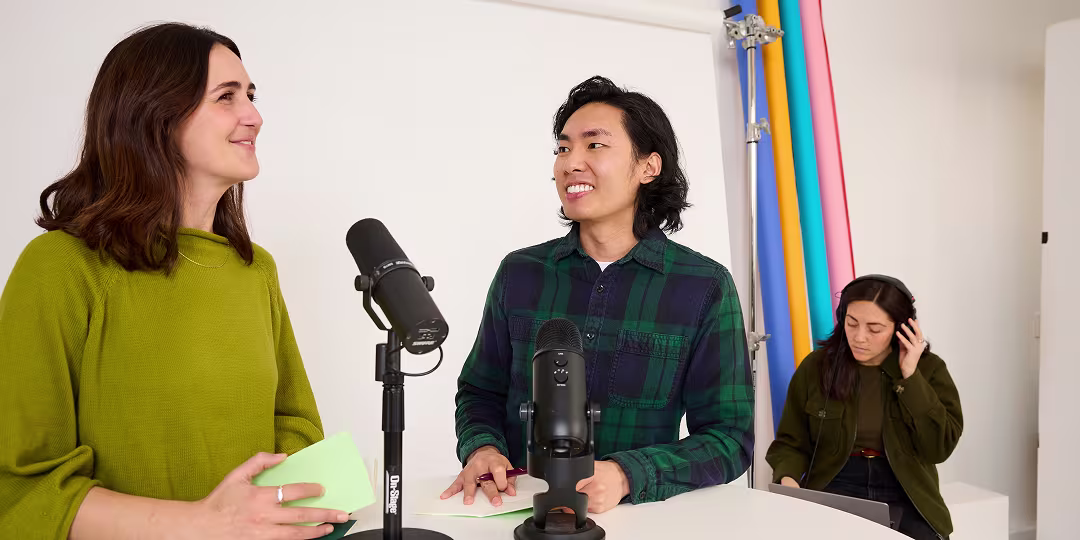 Three people recording a podcast in a bright studio: two speaking into microphones at a table while a third listens on headphones.