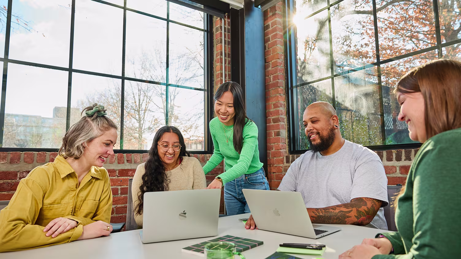 Five marketing professionals gathered around a table with laptops in a sunlit, brick-walled office, smiling and collaborating.