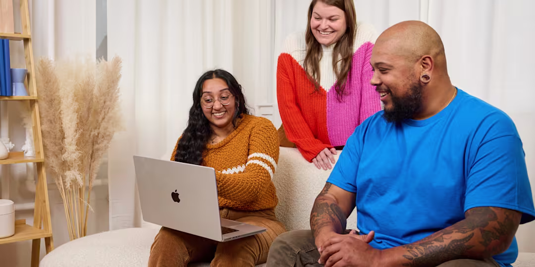 Three people sitting on a couch, laughing and working together. One has a laptop.