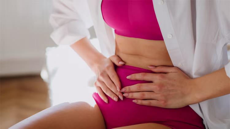Close-up of a woman in a pink sports bra and white shirt, holding her stomach in discomfort.