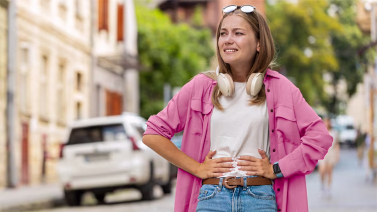 A woman standing on a city street with headphones around her neck, looking uncomfortable and holding her stomach.