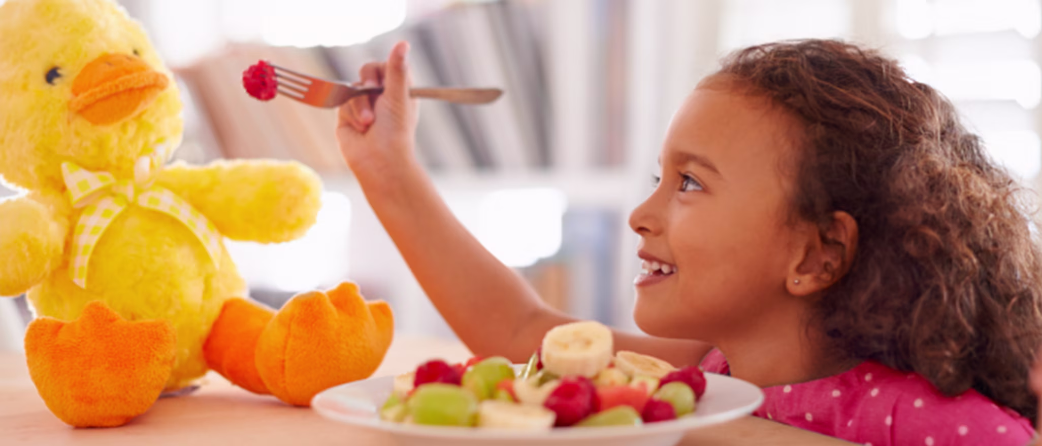 A young girl smiling and feeding a piece of fruit to a yellow plush duck toy.