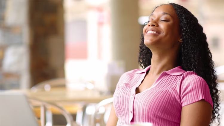 A woman sitting outdoors, closing her eyes and smiling with relief as she enjoys the sun.
