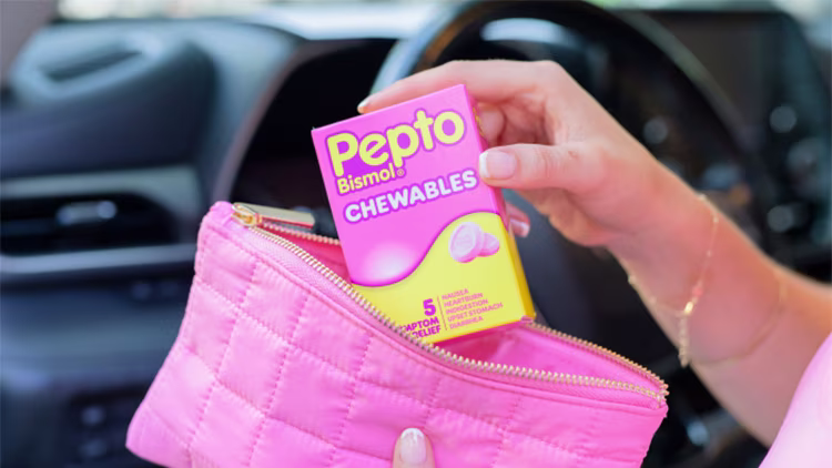 A hand placing a box of Pepto Bismol Chewables into a pink quilted makeup bag inside a car.