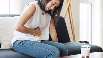 A women sitting on a couch in white top holding stomach in pain and a glass of milk on the table in front of her.