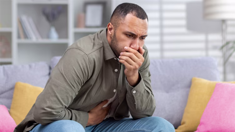 A man sitting on a sofa, looking nauseous with his hand over his mouth and the other on his stomach.