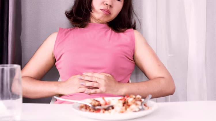 A woman sitting at a table in front of a half-eaten meal, looking bloated and resting her hands on her stomach.