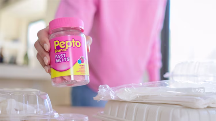 A hand holding a bottle of Pepto Bismol Fast Melts in front of a blurry kitchen background with food containers.