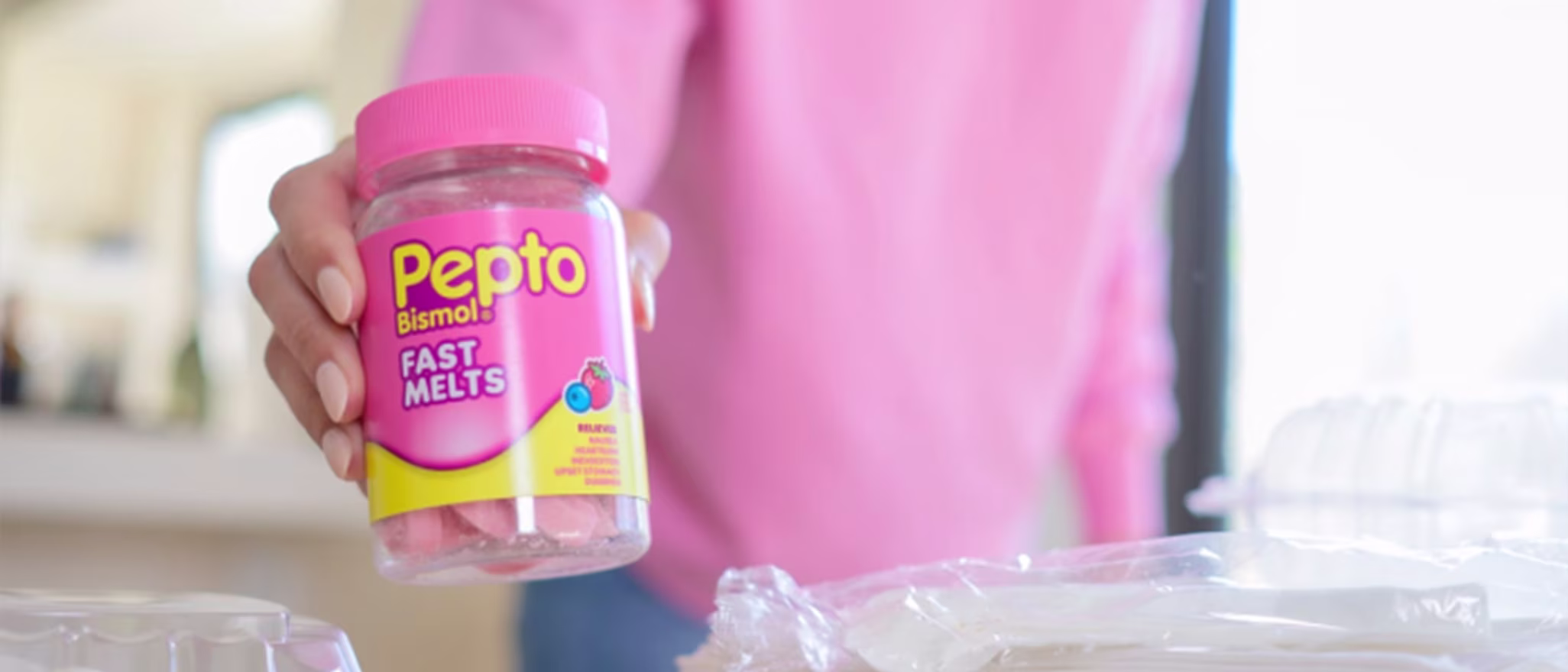 A hand holding a bottle of Pepto Bismol Fast Melts in front of a blurry kitchen background with food containers.