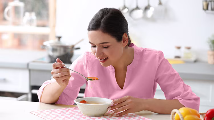 A woman in a pink shirt sitting at a kitchen table, happily eating a bowl of vegetable soup.