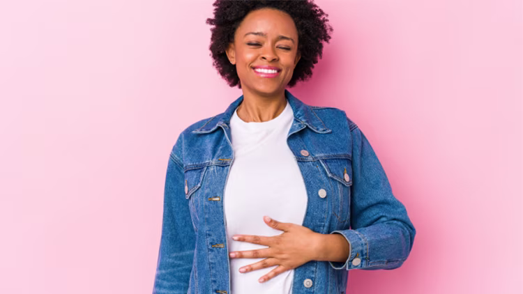A woman in a denim jacket standing against a pink background, smiling and resting her hand on her stomach.