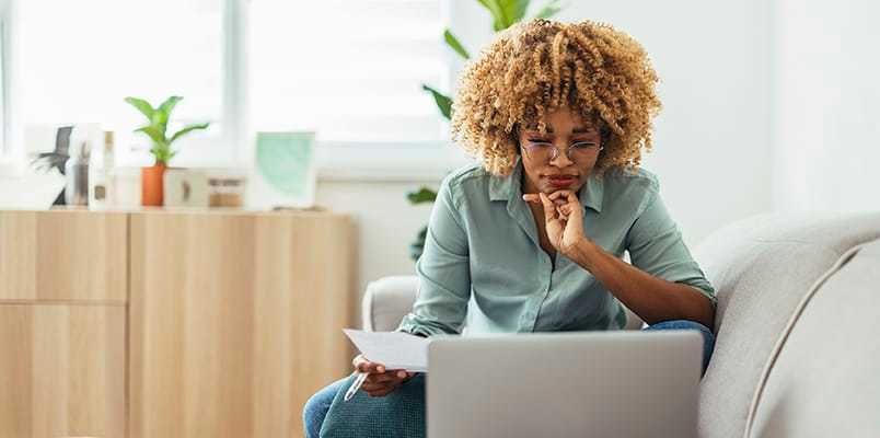 Woman in a blue shirt looks thoughtfully at her laptop while sitting on a couch.