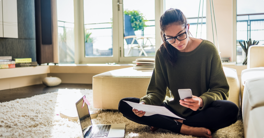 A young woman multitasking, holding tax papers and phone while preparing taxes on her computer