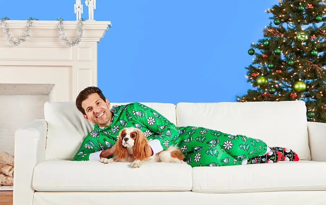 A man in a green Hidden Valley Ranch onesie lies on a white couch with a dog, fireplace and Christmas tree in the background.