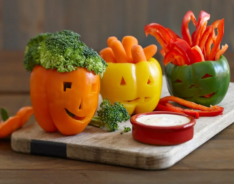 Orange, yellow and green bell peppers carved to look like Jack-o-lanterns on a wood cutting board with dipping vegetables and ranch.