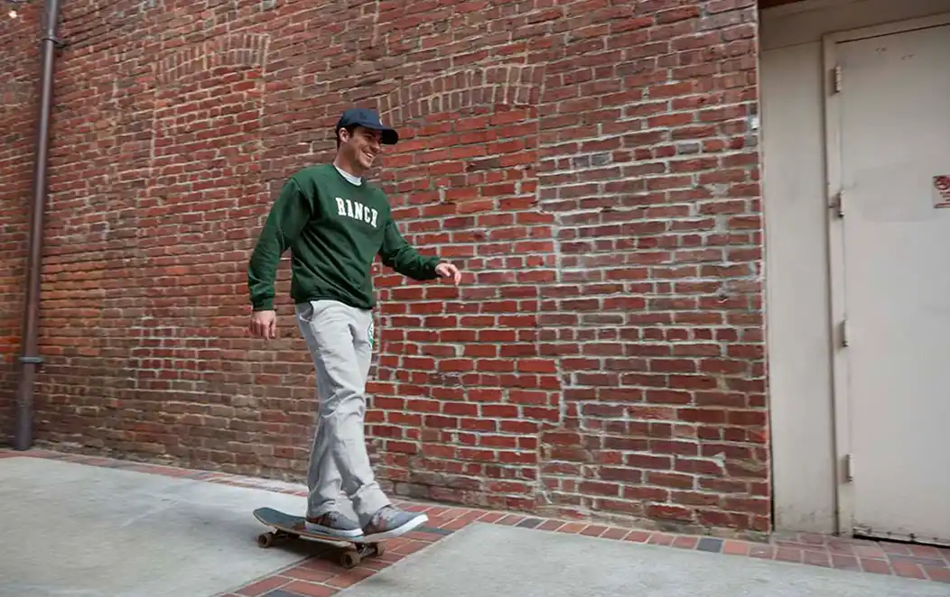 Man in a green sweatshirt with the word Ranch in white letters rides a skateboard next to a brick wall