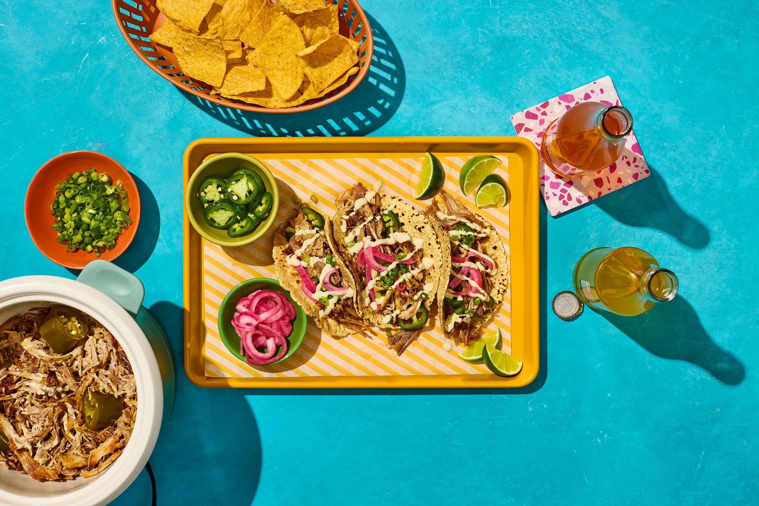 Overhead shot of tray of pulled pork tacos, slow cooker filled with pulled pork, on a turquoise background