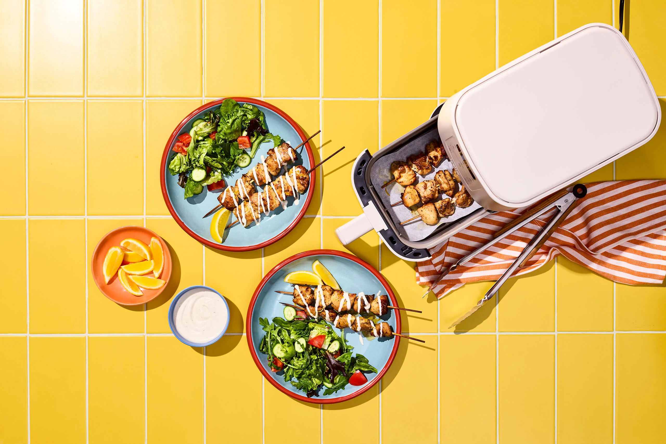 Chicken skewers on plates and in an air fryer, on a yellow tile background