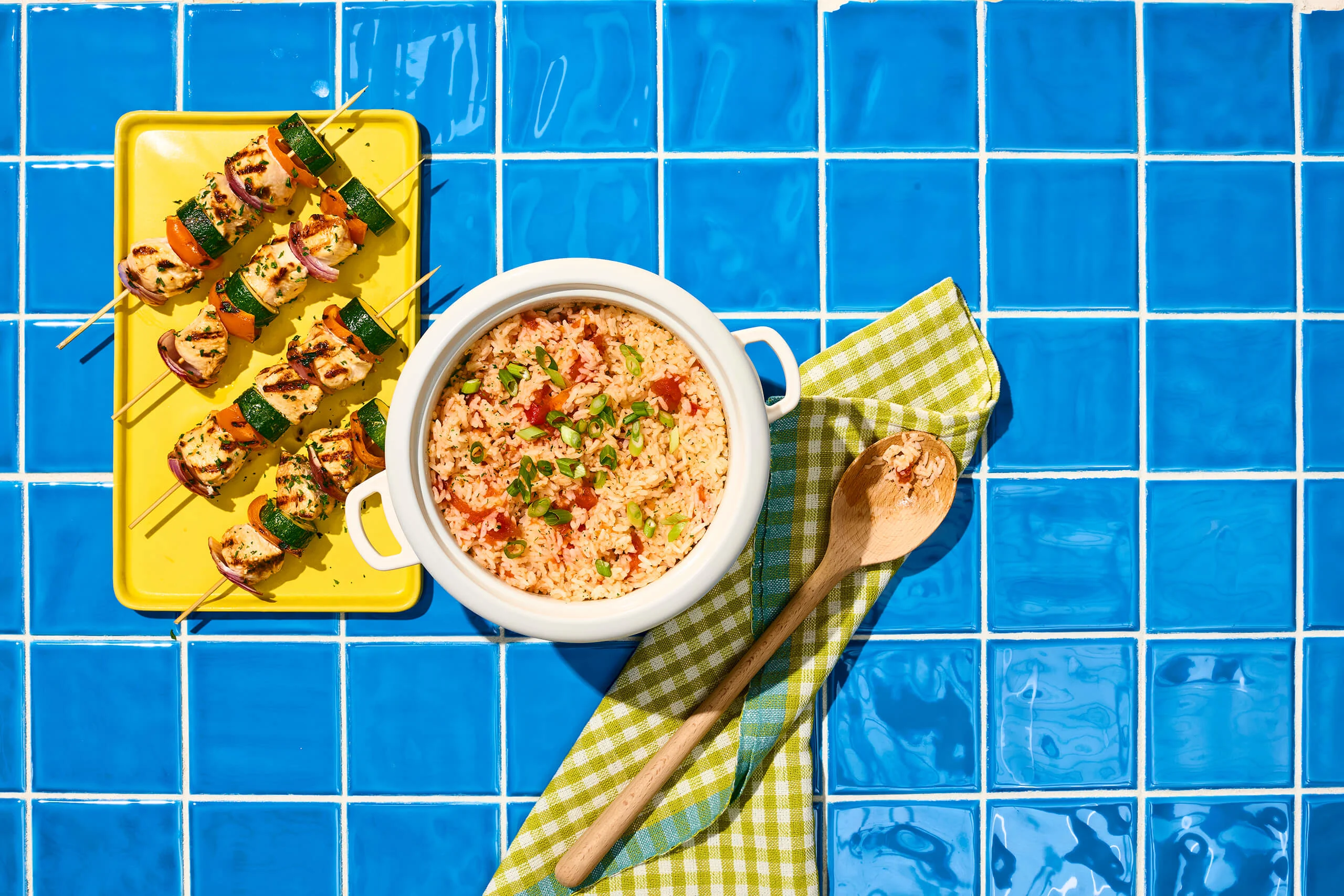 Tomate Rice in a rice cooker, chicken skewers on a yellow platter, blue checked tablecloth background