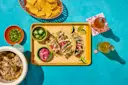 Mexican tacos with shredded meat, pickled onions, jalapenos, lime wedges, and chips on a blue background.