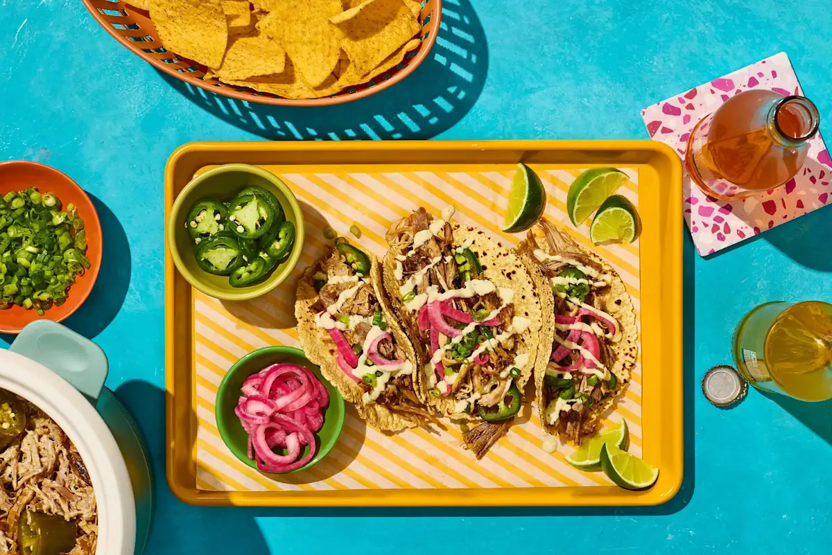 Mexican tacos with shredded meat, pickled onions, jalapenos, lime wedges, and chips on a blue background.