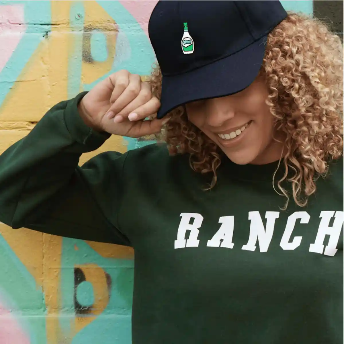 Woman wearing a green sweatshirt with the word "Ranch" on it and a black cap with an embroidered ranch bottle