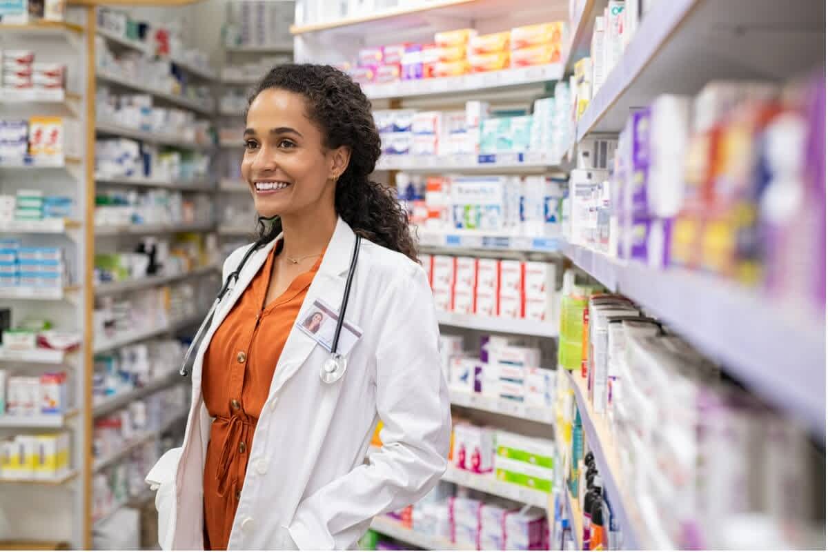 A young lady smiles in white doctor scrubs at her pharmacy